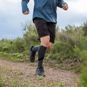 Person running on a trail wearing blue shirt, black shorts, and Injinji Trail Midweight Over the Calf Performance Toe Socks in Granite. Gray with black accents. Anatomical fit toe socks promotes natural toe splay resulting in total foot utilization, which supports full-foot alignment and engagement. Buy now at ICEGRIPPER.