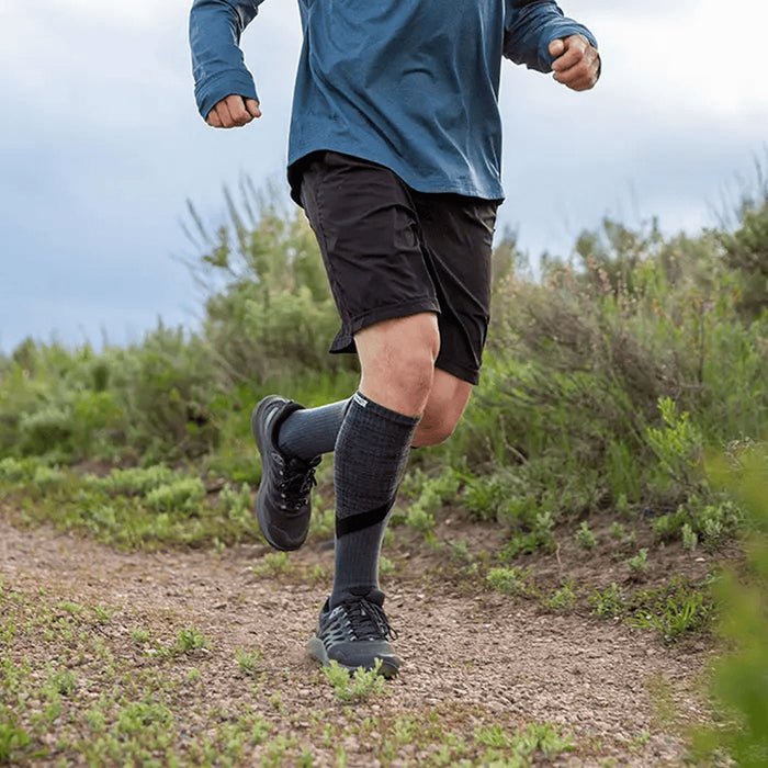 Person running on a trail wearing blue shirt, black shorts, and Injinji Trail Midweight Over the Calf Performance Toe Socks in Granite. Gray with black accents. Anatomical fit toe socks promotes natural toe splay resulting in total foot utilization, which supports full-foot alignment and engagement. Buy now at ICEGRIPPER.