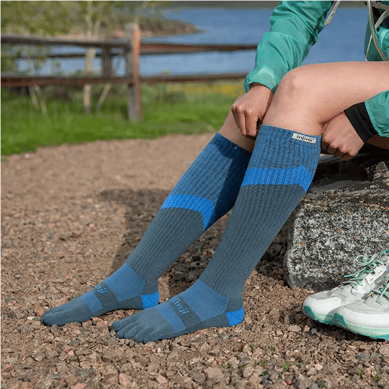 Person wearing a pair of of Injinji Trail Midweight Over the Calf – Performance Toe Socks in Storm -  blue and gray toe-separated socks outdoors by a lake. Buy now at ICEGRIPPER.