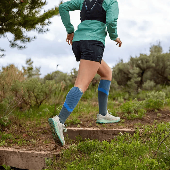 Person running on a trail wearing green shirt, black shorts, and Injinji Trail Midweight Over the Calf Performance Toe Socks in Storm. Blue and gray toe-separated blister prevention socks. Buy now at ICEGRIPPER.