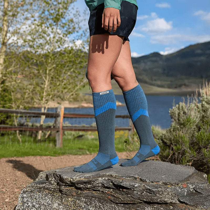 Person wearing a pair of of Injinji Trail Midweight Over the Calf – Performance Toe Socks in Storm -  blue and gray toe-separated socks outdoors by a lake. Buy now at ICEGRIPPER.