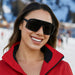 Woman wearing black sunglasses and a red ski jacket with a blurred snowy.  background. The Black on Black Campeone, built around Knockaround’s innovative Knockterra performance lens technology, Campeone sunglasses deliver crystal-clear vision, UV400 protection, and impact-resistant durability. Their sleek wraparound design guards against low winter sun and glare reflected from snow and ice, while rubberized adjustable nose grips and flexible non-slip arms ensure a secure, all-day fit during high-energy wint