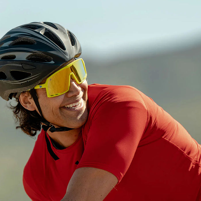 Person wearing a red cycling outfit with a helmet and yellow sunglasses, outdoors. The High Voltage Campeone is built around Knockaround’s innovative Knockterra performance lens technology, Campeone sunglasses deliver crystal-clear vision, UV400 protection, and impact-resistant durability. Their sleek wraparound design guards against low winter sun and glare reflected from snow and ice, while rubberized adjustable nose grips and flexible non-slip arms ensure a secure, all-day fit.