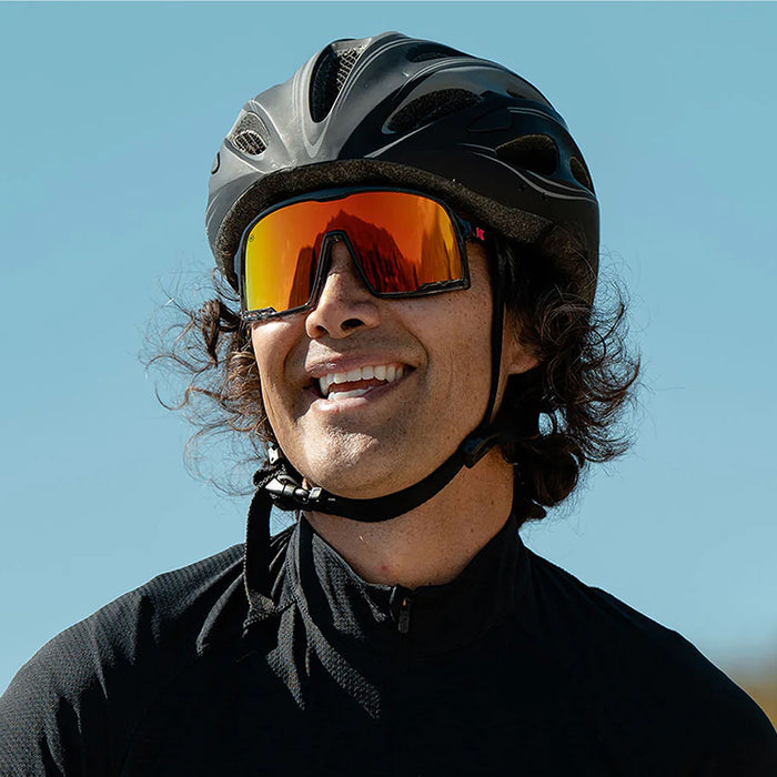 Person wearing a black helmet and reflective sunglasses against a clear blue sky. The Magma Campeone is built around Knockaround’s innovative Knockterra performance lens technology, Campeone sunglasses deliver crystal-clear vision, UV400 protection, and impact-resistant durability. Their sleek wraparound design guards against low winter sun and glare reflected from snow and ice, while rubberized adjustable nose grips and flexible non-slip arms ensure a secure, all-day fit.