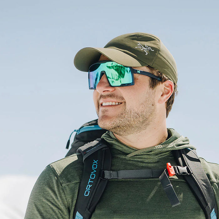 Man wearing a green cap and sunglasses with a backpack, standing against a light blue sky. The Navy Mint Campeone is built around Knockaround’s innovative Knockterra performance lens technology, Campeone sunglasses deliver crystal-clear vision, UV400 protection, and impact-resistant durability. Their sleek wraparound design guards against low winter sun and glare reflected from snow and ice, while rubberized adjustable nose grips and flexible non-slip arms ensure a secure, all-day fit during high-energy.
