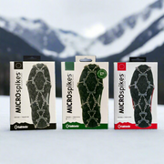 Pack shot of all 3 Kahtoola Microspike variants: Black, Green and Red. The packs are sat in the snow at the base of a mountain.