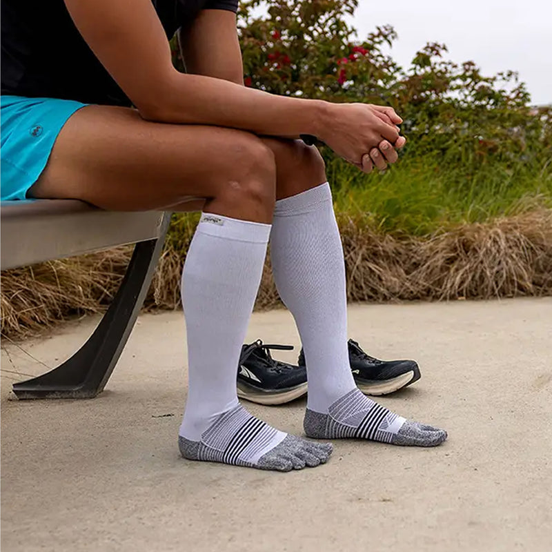 A person sitting on a bench outdoors wearing Injinji Compression Over the Calf Toe Socks. White and Grey ribbed toe socks with gray striped toes, paired with black athletic shoes. The background shows a concrete surface, grass, and shrubs. Buy now at ICEGRIPPER