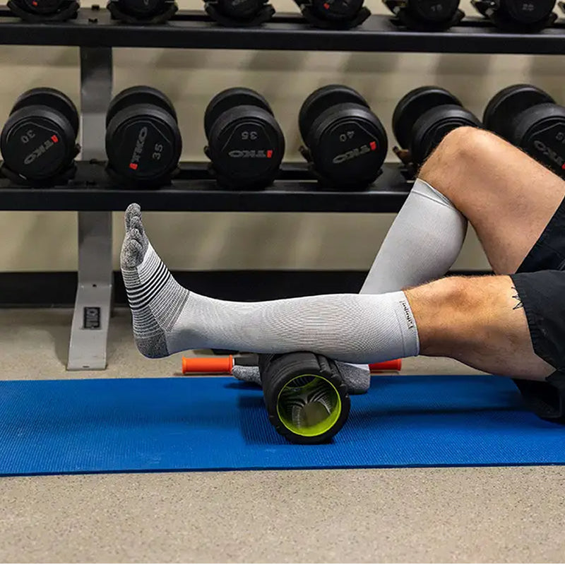 Injinji Compression Over the Calf Toe Socks. Buy now at ICEGRIPPER. Person using a foam roller on a blue mat with gym equipment in the background.
