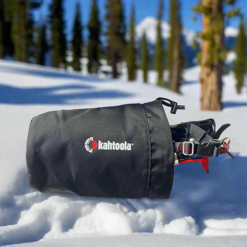 Black Kahtoola KTS Tote, with Kahtoola KTS Crampons stowed inside. The bag is sat on a snowy mountainside with alpine trees behind, demonstrating the type of environment where the bag would typically be used. Buy now at ICEGRIPPER.